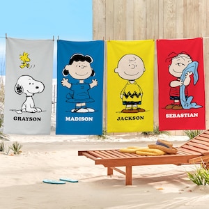 May include: Four colorful beach towels hanging on a clothesline. Each towel features a different Peanuts character: Snoopy, Lucy, Charlie Brown, and Linus. The towels are gray, blue, yellow, and red, with names printed below the characters. A wooden beach chair sits in the foreground.
