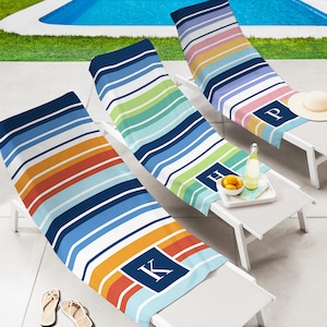May include: Three striped beach towels with personalized initials, "K", "H", and "P", are laid out on white lounge chairs. The chairs are positioned near a swimming pool.