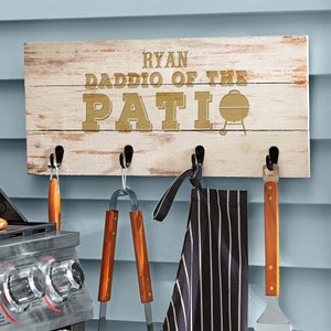 May include: A white wooden sign with the text "Ryan Daddo of the Patio" in gold lettering. The sign has five hooks for hanging items, and a black and white striped apron, a spatula, and tongs are hanging from the hooks.