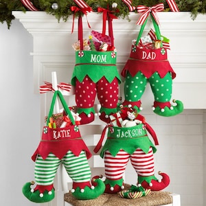 May include: Four Christmas elf-themed gift bags hanging from a white mantel. Each bag is red and green, shaped like elf legs, and filled with gifts. The bags are labeled "MOM", "DAD", "KATIE", and "JACKSON".