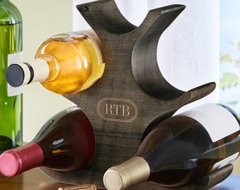 May include: A wooden wine rack with three bottles of wine. The rack is dark brown with a curved design and holds the bottles horizontally. The initials "RTB" are engraved on the front. A corkscrew and cork are in the foreground.