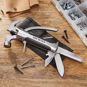 May include: A multi-tool with a hammer, pliers, knife, and saw. The tool is open and has a black handle with the name "MICHAEL" engraved on it. The tool is on a wooden surface with nails and screws.