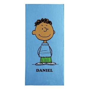 May include: A light blue rectangular towel featuring a cartoon illustration of a boy with brown skin, black curly hair, and a smiling face. He wears a blue shirt and green pants. The name "DANIEL" is printed in black at the bottom.