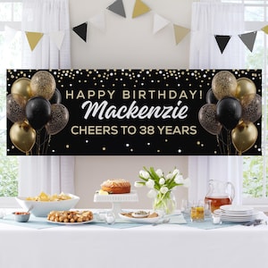 May include: A black banner with gold confetti, balloons, and the text "HAPPY BIRTHDAY! Mackenzie CHEERS TO 38 YEARS." The banner is decorated with gold, black, and glitter balloons. A table is set with food and drinks.