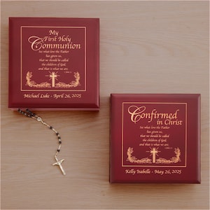 May include: Two red wooden boxes with gold lettering. The top box has the text "My First Holy Communion" and the bottom box has the text "Confirmed in Christ". Both boxes have a cross and floral design. The boxes are on a wooden surface.