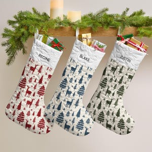 May include: Three quilted Christmas stockings featuring a white cuff with the names REGINE, BLAKE, and JUSTIN. The stockings have a repeating pattern of deer and pine trees in red, blue, and green, each topped with small wrapped gift boxes.