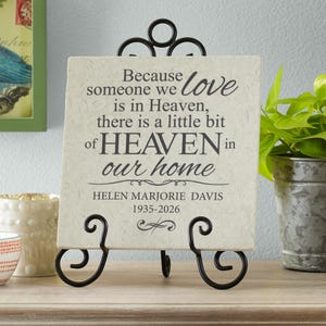 May include: A memorial plaque with a heartfelt message, displayed on a decorative black metal stand. The plaque features the text "Because someone we love is in Heaven, there is a little bit of HEAVEN in our home."