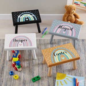 May include: Four small wooden step stools with rainbow designs and personalized names. The stools are painted black, white, gray, and tan. Each stool has a different name: Brighton, Harper, Abigail, and Carter. The stools are on a patterned rug.
