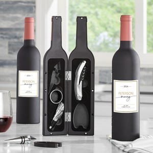 May include: A black wine bottle with a pink top and a white label with gold trim. A wine bottle-shaped case holds wine tools, including a corkscrew and a bottle stopper. The label reads "Peterson Winery".