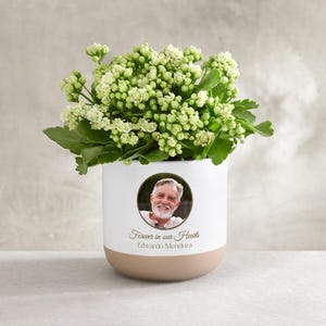 May include: A white and tan ceramic planter with a photo of a smiling man. The planter is filled with green plants and white flowers. Text reads "Forever in our Hearts" and the name "Eduardo Mendoza".