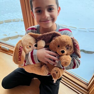 May include: A young boy wearing a striped shirt is holding two plush brown dogs. One dog has the word "Mommy" embroidered on it and the other has the word "Card" embroidered on it.