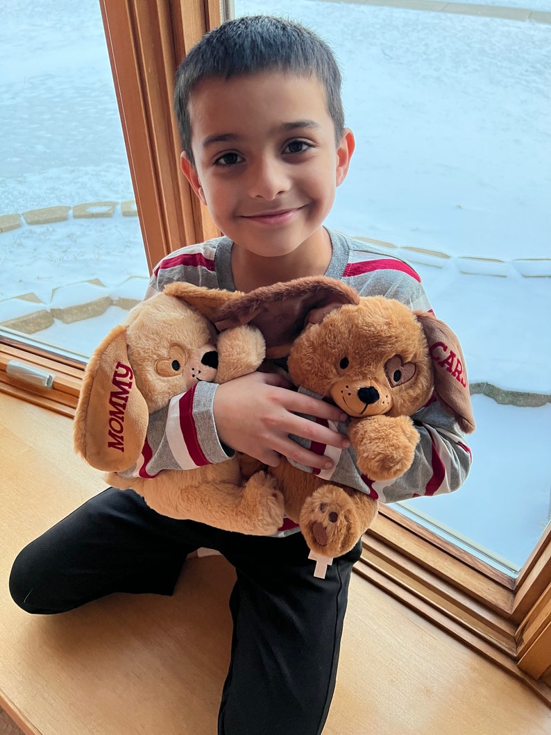 May include: A young boy wearing a striped shirt is holding two plush brown dogs. One dog has the word "Mommy" embroidered on it and the other has the word "Card" embroidered on it.