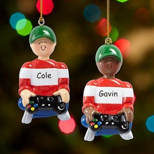 May include: Two personalized Christmas ornaments of boys wearing red and white striped shirts, green hats, and blue jeans. They are holding video game controllers. The ornaments are labeled "Cole" and "Gavin".