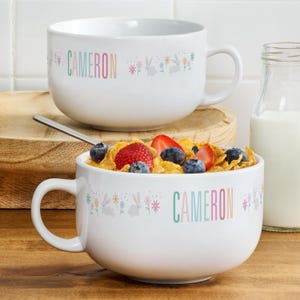 May include: Two white ceramic bowls with handles, one filled with cereal, strawberries, and blueberries. The bowls have the name "CAMERON" in pastel colors and a floral and bunny design. A spoon rests in the bowl.