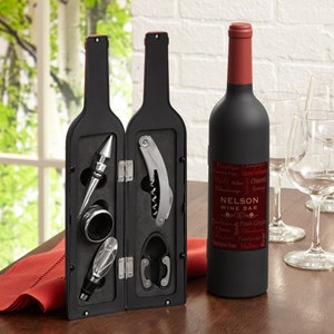 May include: A black wine bottle shaped box containing a set of wine tools. The tools include a bottle opener, a corkscrew, a bottle stopper, and a foil cutter. The box is open and the tools are visible. The box is on a wooden table with a red napkin.