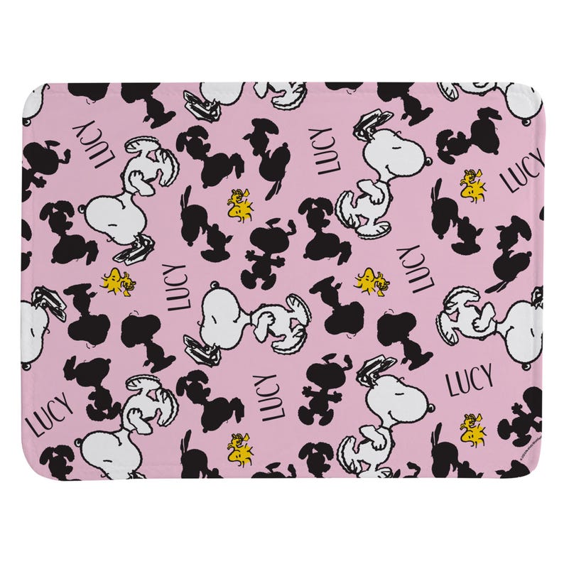 May include: A rectangular bath mat with a light pink background featuring a repeating pattern of white Snoopy characters, black Snoopy silhouettes, small yellow Woodstock figures, and the name Lucy printed in a black sans-serif font throughout.
