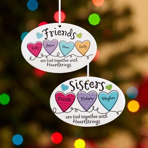 May include: Two oval-shaped ceramic ornaments with colorful hearts and the words "Friends" and "Sisters" with names written inside the hearts. The text reads "are tied together with Heartstrings".