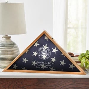 May include: A wooden flag display case in a triangular shape, showcasing a folded American flag with white stars on a blue field. The case includes a printed inscription with a name and text.