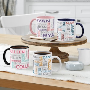 May include: Four white ceramic mugs with personalized names in various colours. One mug has a pink handle and rim, another has a navy blue handle and rim, a third has a black handle and rim, and the last has a white handle. The mugs are on a wooden cake stand.