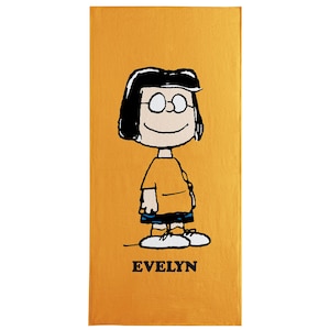 May include: A rectangular, mustard-colored towel featuring a cartoon illustration of a character with black hair, glasses, and an orange shirt. The character is standing, with the name "EVELYN" printed in black at the bottom.