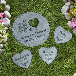 May include: Four gray stone heart-shaped memorial garden stones with engraved messages. The largest stone reads "Always & Forever In Our Hearts". The other stones read "Grandma Lucy 1927-2010", "Uncle Jim 1950-2019", and "Shadow 2012-2025".