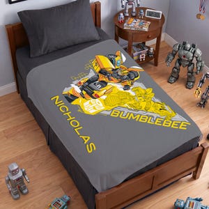 May include: A gray fleece blanket featuring a yellow Bumblebee character graphic, the Transformers logo, and the name NICHOLAS printed in yellow text. The design includes a dynamic illustration of the robot in both vehicle and robot forms.