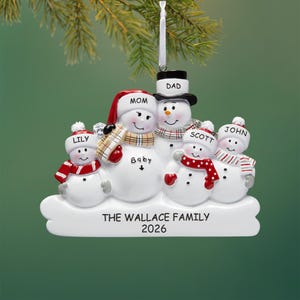 Peut inclure: Une décoration de Noël représentant une famille de cinq bonhommes de neige sur un socle blanc portant l'inscription THE WALLACE FAMILY 2026. Les personnages sont nommés DAD, MOM, BABY, LILY, SCOTT et JOHN, avec des écharpes et bonnets colorés.