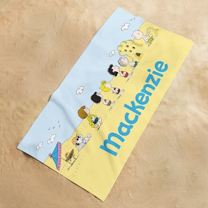 May include: A beach towel featuring a cartoon beach scene with characters and the name "Mackenzie" in blue. The towel has a yellow and light blue color scheme, with cartoon characters enjoying the beach.