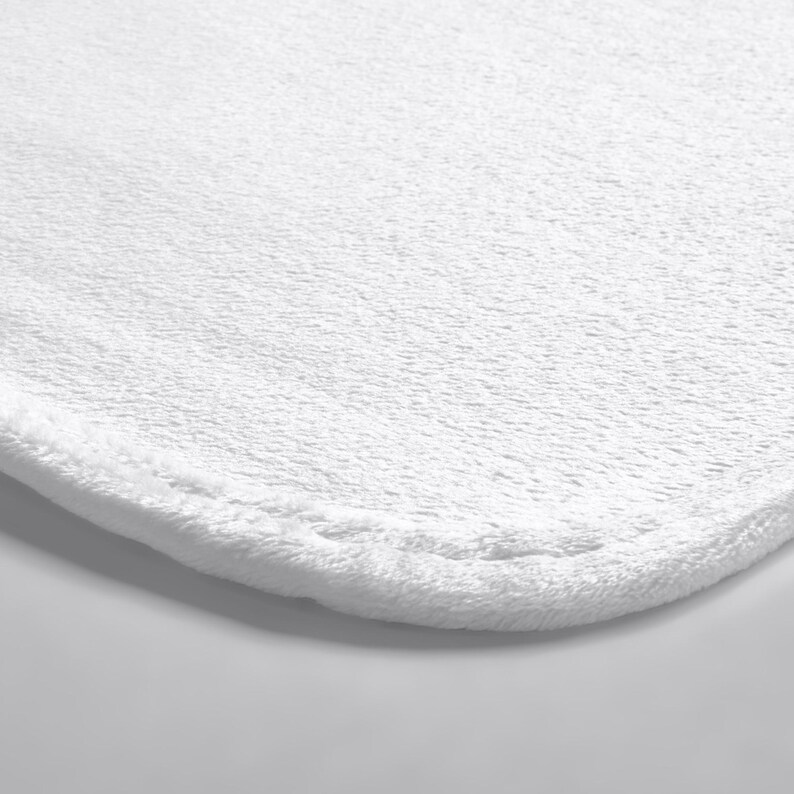 May include: A solid white bath mat featuring a soft, plush, and textured fabric surface with rounded corners and a stitched hem along the edge. The material has a dense, short-pile construction that creates a uniform and clean appearance.