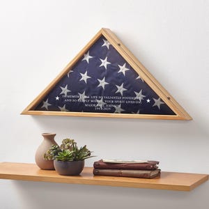 May include: A wooden triangular flag display case holding a folded American flag with white stars on a blue field. The case has a glass front and includes text: "IN MEMORY OF A LIFE SO VALIANTLY FOUGHT FOR...MAJOR PAUL JOHNSON 1947-2025."