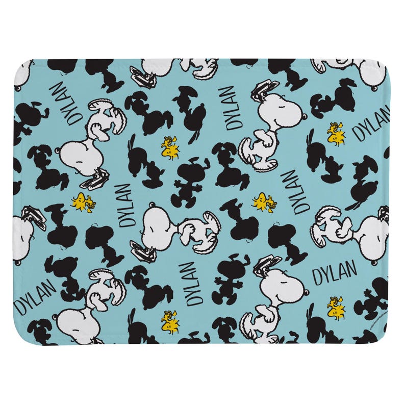 May include: A light blue rectangular mat featuring a repeating pattern of white Snoopy characters, black Snoopy silhouettes, small yellow Woodstock figures, and the name DYLAN printed in black text throughout the design on the surface.