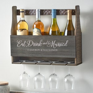 May include: A rustic wooden wall-mounted wine rack with a gray finish. It features a sign that reads "Eat, Drink and be Married" with the names Cameron & Mackenzie. The rack holds four bottles of wine and has four glass holders underneath.