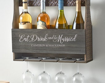 Puede incluir: Un estante de vino de pared de madera rústica con un acabado gris. Cuenta con un letrero que dice "Eat, Drink and be Married" con los nombres Cameron & Mackenzie. El estante tiene capacidad para cuatro botellas de vino y tiene cuatro soportes para vasos debajo.