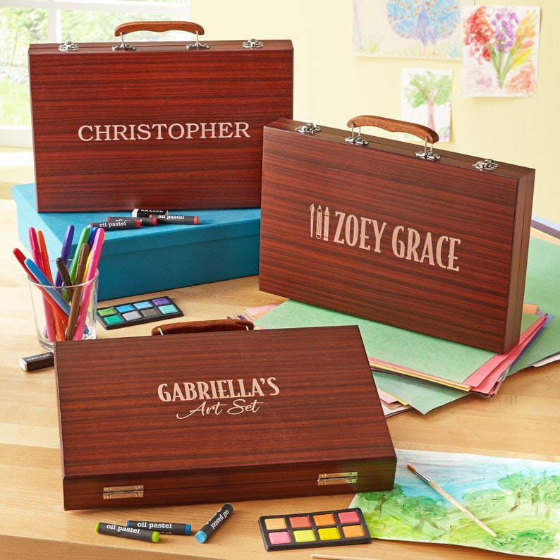 May include: Three wooden art cases with personalized names. The cases are brown with a wood grain pattern. The top case says "CHRISTOPHER", the middle case says "ZOEY GRACE", and the bottom case says "GABRIELLA'S Art Set".