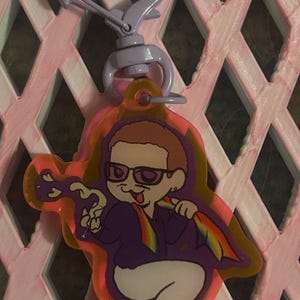 May include: A novelty keychain featuring a cartoon character with glasses, a purple suit, and a rainbow tie. The keychain has a purple clasp and a pink and orange border. The background is a pink lattice.
