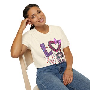 Puede incluir: Camiseta color crema con la palabra "LOVE" en un diseño decorativo. La "L" es morada con patrones de corazones, la "O" tiene forma de corazón con un lazo rosa, la "V" tiene corazones rojos y la "E" tiene estampado de leopardo.