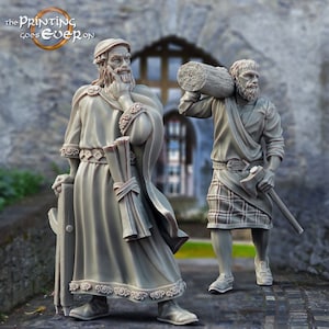 Könnte beinhalten: Zwei 3D-gedruckte Figuren, eine von einem Mann in einem langen Gewand, der eine Schriftrolle und einen Stab hält, und die andere von einem Mann, der einen Baumstamm auf der Schulter trägt und eine Axt hält. Die Figuren stehen auf einem Steinweg vor einer Steinmauer mit einem Fenster.