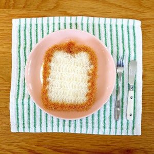 May include: A crocheted bread slice in a light pink bowl on a white and green striped towel. The bread is brown on the outside and white on the inside. There is a silver fork and a silver butter knife next to the bowl.