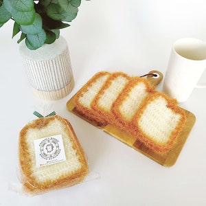 May include: Four crocheted bread slices in orange and white, arranged on a wooden cutting board. The bread slices are made to look like real bread. The text "Freshly Baked Bread Scrub" is printed on a white label.