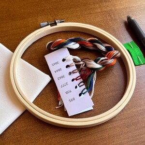 Shows the supplies included in this Halloween craft kit, including a bamboo embroidery hoop, a set of labeled DMC embroidery floss, cream colored cotton fabric, a heat erase pen used to transfer the pattern to fabric, an embroidery needle and the pattern with detailed instructions on how to complete the craft project.