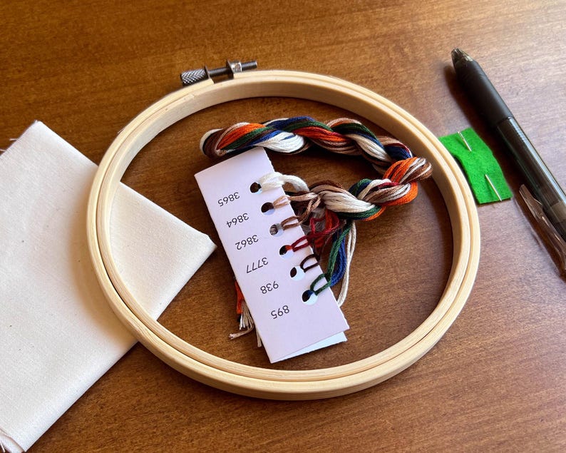 Shows the supplies included in this Halloween craft kit, including a bamboo embroidery hoop, a set of labeled DMC embroidery floss, cream colored cotton fabric, a heat erase pen used to transfer the pattern to fabric, an embroidery needle and the pattern with detailed instructions on how to complete the craft project.