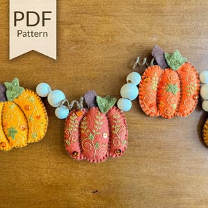 Pode incluir: Uma guirlanda de abóboras de feltro em tons de laranja e vermelho, decoradas com vinhas bordadas em verde e pequenas lantejoulas. As abóboras estão enfiadas num cordão com contas de madeira branca. Uma etiqueta PDF Pattern é visível.