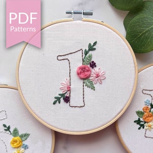 May include: Three embroidery hoops with floral designs and numbers 1, 2, and 5. The hoops are made of wood and have a white fabric background. The flowers are embroidered in pink, yellow, orange, and green thread.