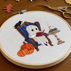 Finished embroidered hoop art with a cute ghost reading a book in a blue armchair with a crow perched on the back. There are pumpkins in the foreground and a side table with a cup of coffee and books on it.