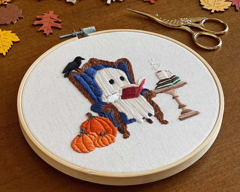 Finished embroidered hoop art with a cute ghost reading a book in a blue armchair with a crow perched on the back. There are pumpkins in the foreground and a side table with a cup of coffee and books on it.