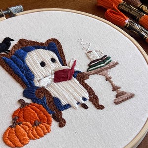 Close up detail showing the satin stitch in this hand embroidered artwork of a ghost sitting in a blue armchair reading a book. The ghost is white with black eyes. There is a black bird perched on the back of the chair. A small table with a cup and saucer sits next to the chair. There are pumpkins in the foreground.