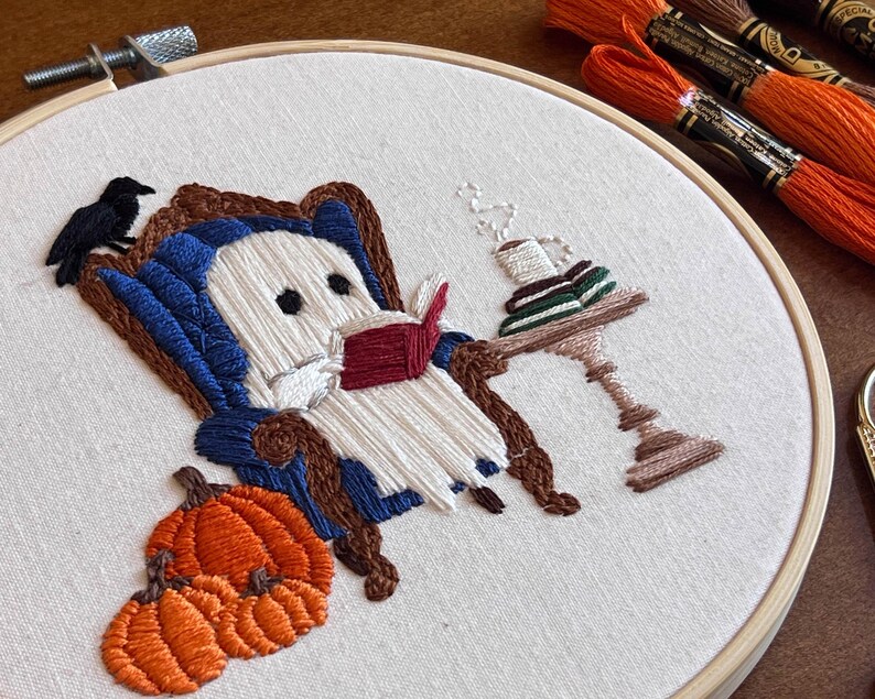 Close up detail showing the satin stitch in this hand embroidered artwork of a ghost sitting in a blue armchair reading a book. The ghost is white with black eyes. There is a black bird perched on the back of the chair. A small table with a cup and saucer sits next to the chair. There are pumpkins in the foreground.