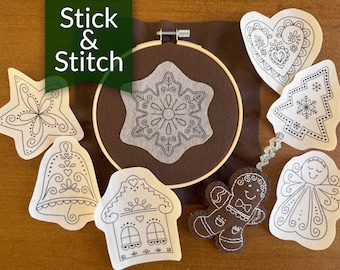 May include: A set of eight white felt ornaments with black embroidery designs. The ornaments include a snowflake, a bell, a star, a gingerbread person, a house, a heart, a Christmas tree, and an angel. The ornaments are arranged on a brown felt background with the words "Stick & Stitch" in green.