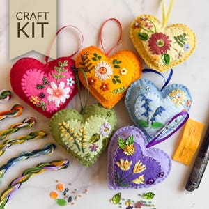 May include: A craft kit featuring heart-shaped ornaments in various colors, each adorned with embroidered floral designs. The hearts are red, orange, yellow, blue, green, and purple. The kit includes embroidery floss, needles, and embellishments. The text "CRAFT KIT" is visible.