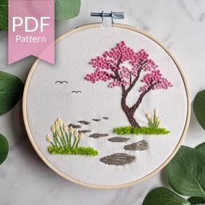 May include: Embroidered hoop art featuring a pink and white cherry blossom tree with a brown trunk, green grass, and grey stone stepping stones. The hoop is white with a wooden frame.
